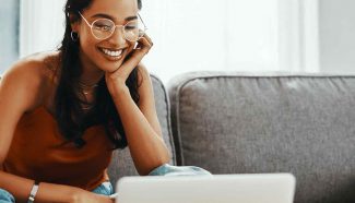 A woman wearing circular glasses smiles as she reviews egg donor requirements on her laptop from her couch.