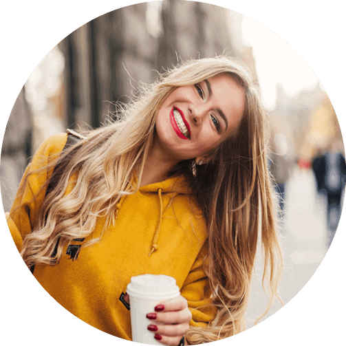 A woman in a yellow sweatshirt holding coffee smiles and appears to be traveling abroad.