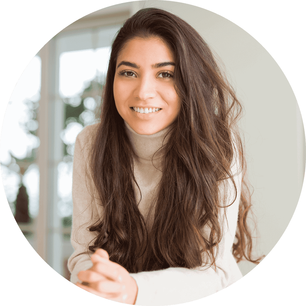 A woman wearing a taupe turtleneck is smiling and poses with her hands clasped leaning against a countertop.