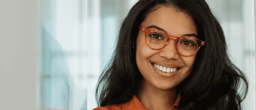 A woman in trendy orange glasses and a matching button-up shirt is smiling in an bright, clean setting.