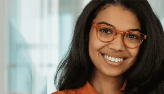 A woman in trendy orange glasses and a matching button-up shirt is smiling in an bright, clean setting.