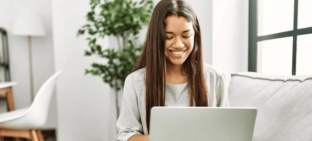 A woman wearing a light gray sweatshirt sits on her couch and browses the internet on her laptop from her couch.