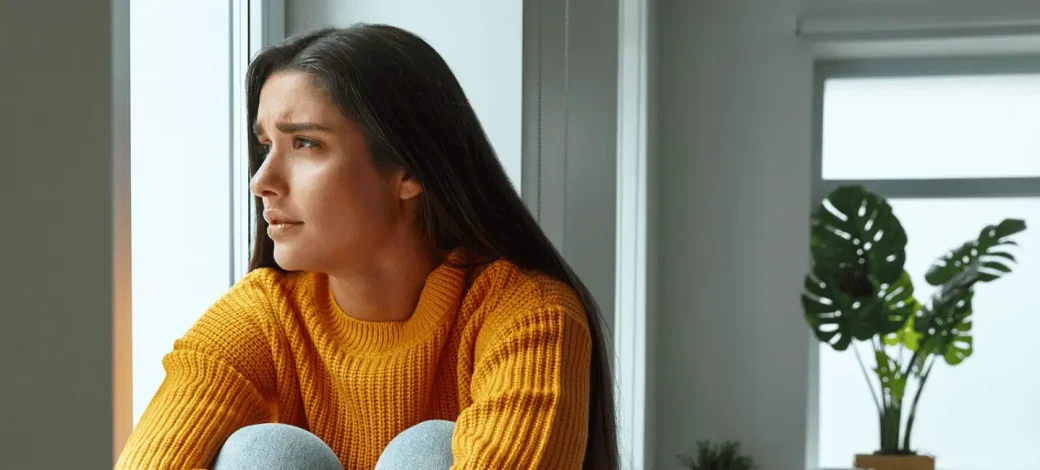 A woman in a bright orange sweater looks out the window with a worried expression.